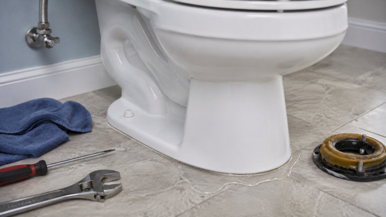 A close-up of a white toilet base next to a replaced toilet flange and wax ring on a tile floor with tools.