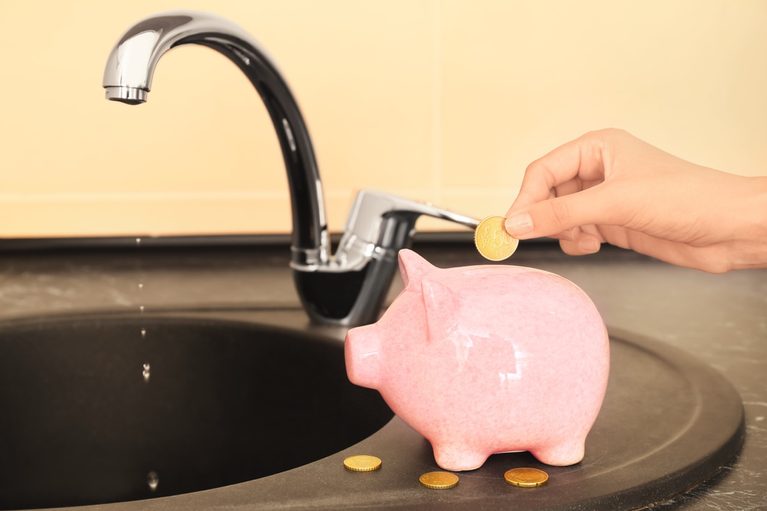Hand dropping a coin into a pink piggy bank under a running water faucet