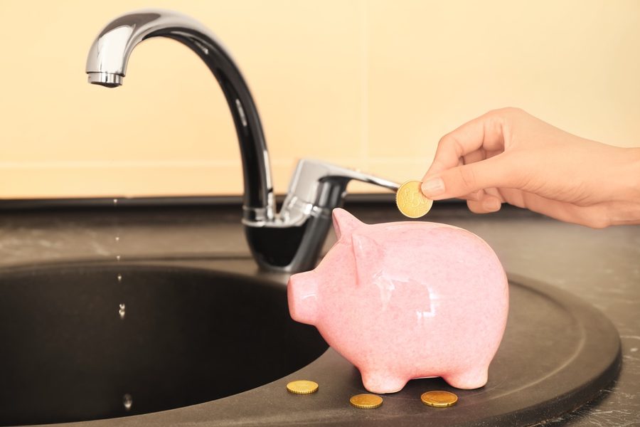 Hand dropping a coin into a pink piggy bank under a running water faucet